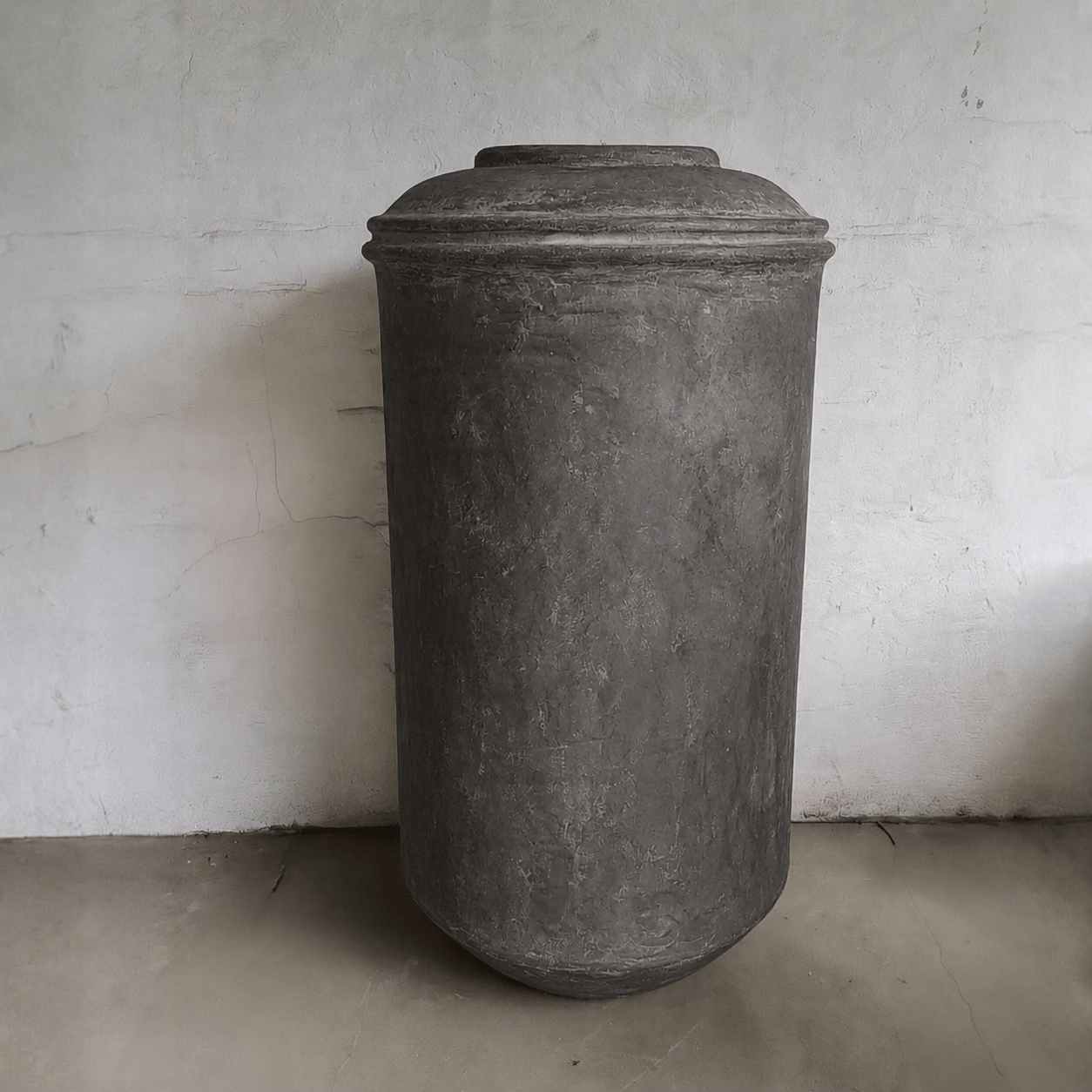 The Damascus Organic Fiberglass Pot - Earth Range by Florastyle By Hingham, a tall cylindrical planter with a round lid, stands against a textured light wall on a smooth floor.
