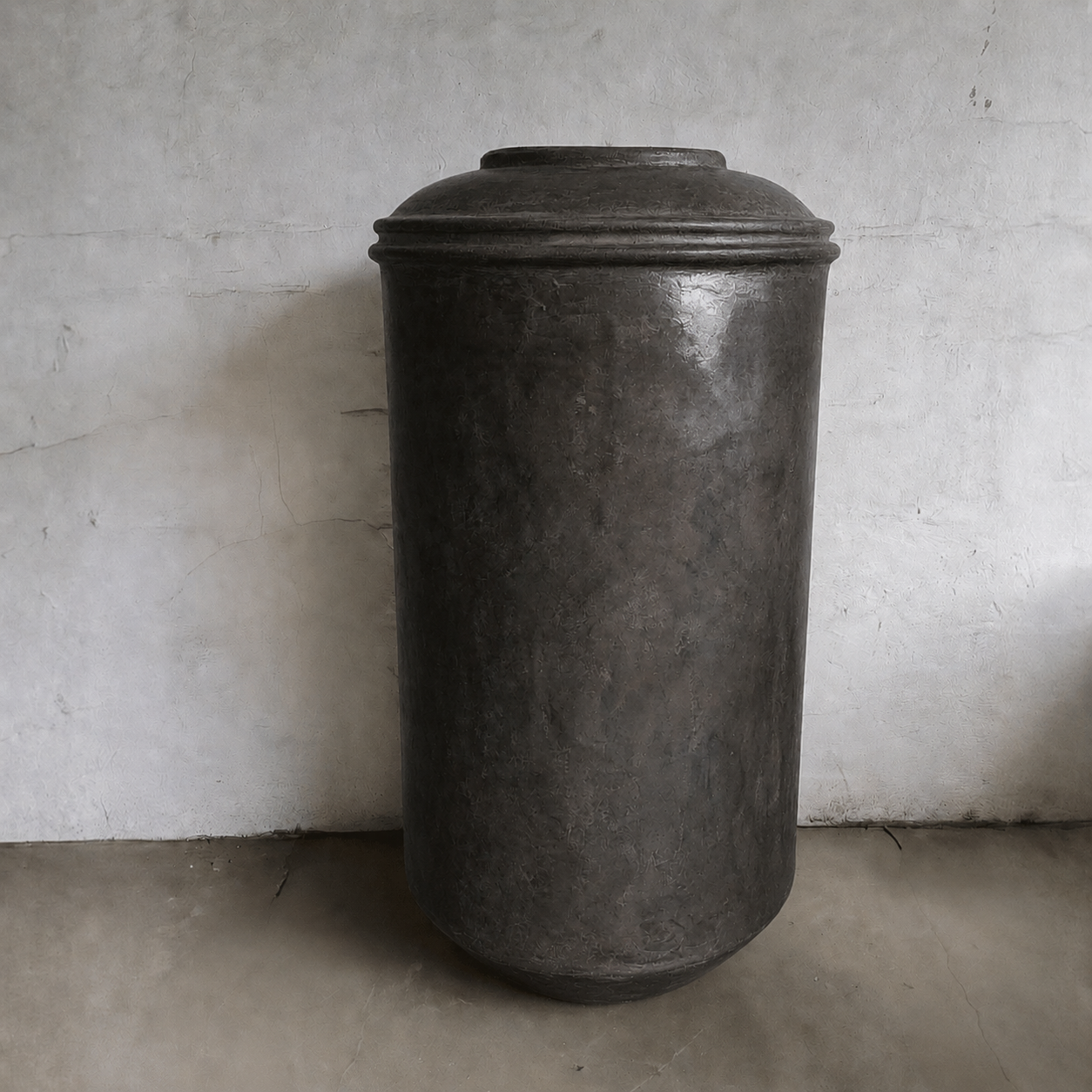 The Damascus Organic Fiberglass Pot - Earth Range by Florastyle By Hingham features a tall cylindrical design with a dark rounded lid, enhancing modern elegance on a plain concrete floor against a textured, light wall.
