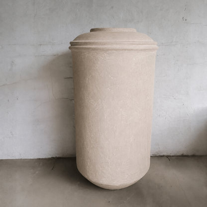 The Florastyle By Hingham Damascus Organic Fiberglass Pot - Earth Range, large and beige with a rounded lid, stands against a plain textured gray wall on a smooth concrete floor.