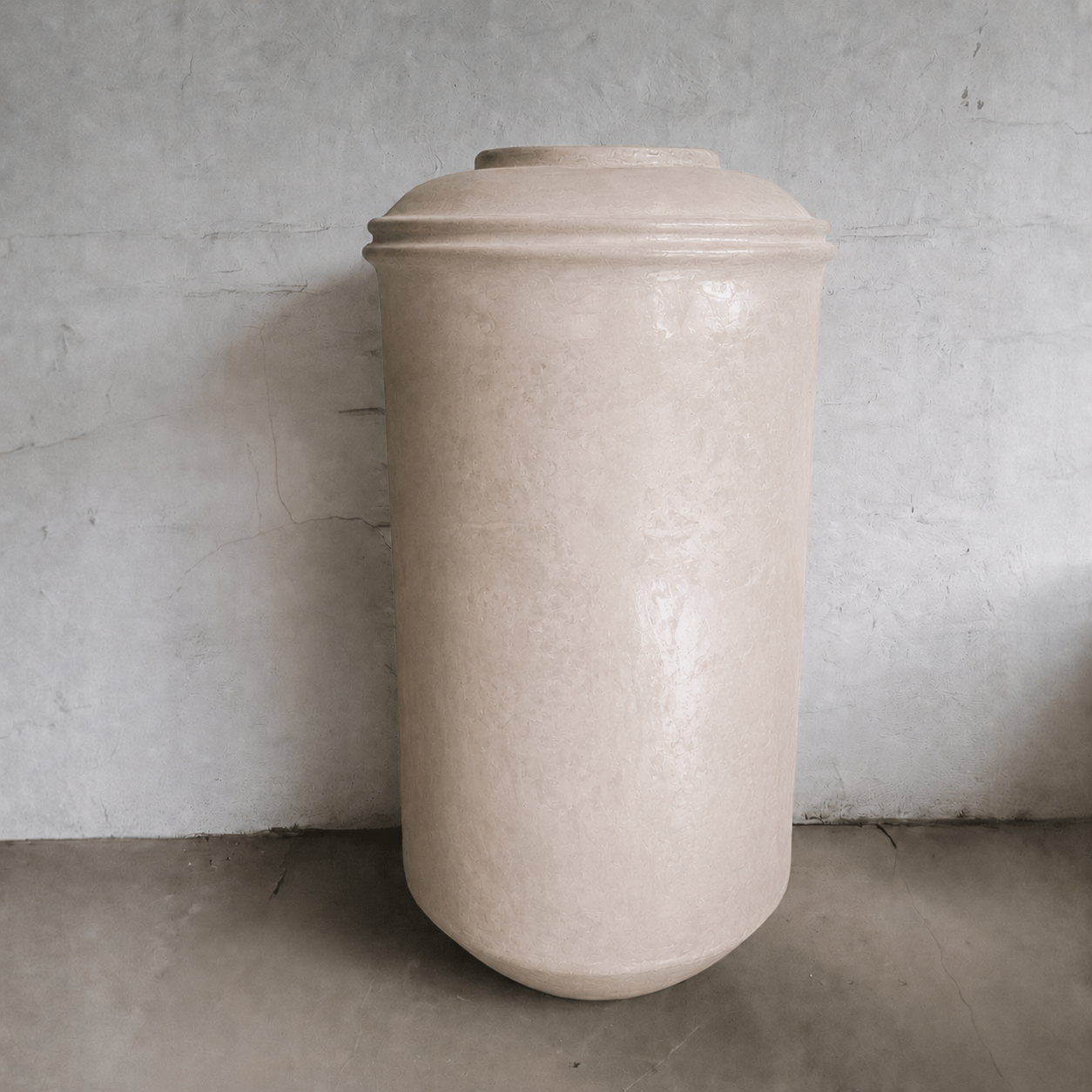 A large, beige Damascus Organic Fiberglass Pot from Florastyle By Hingham’s Earth Range, featuring a wide body and narrow top, rests on a concrete floor against a textured gray wall.