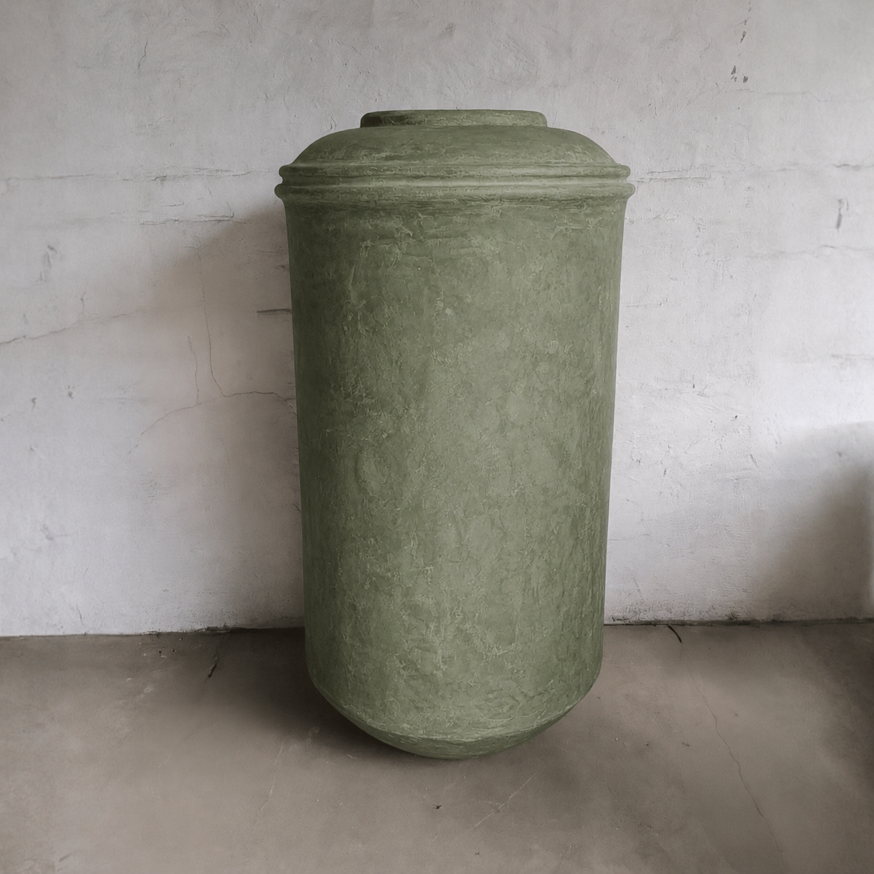 The Damascus Organic Fiberglass Pot – Earth Range by Florastyle By Hingham stands tall and cylindrical with a rounded lid, its green finish contrasting a textured white wall atop a concrete floor.