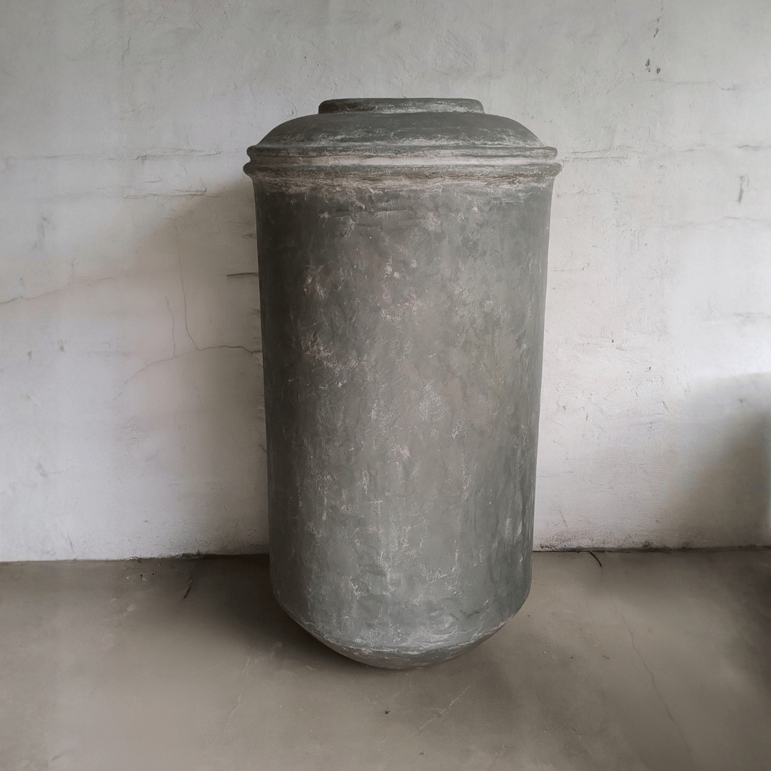 The Damascus Organic Fiberglass Pot - Earth Range by Florastyle By Hingham, a large, tall cylindrical planter in weathered gray, with a fitted lid, stands upright against a textured white wall on a smooth concrete floor.