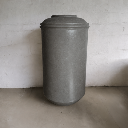 The Florastyle By Hingham Damascus Organic Fiberglass Pot - Earth Range, a tall, cylindrical planter with a rounded base and domed lid, sits against a plain concrete wall and floor—ideal for modern interiors.