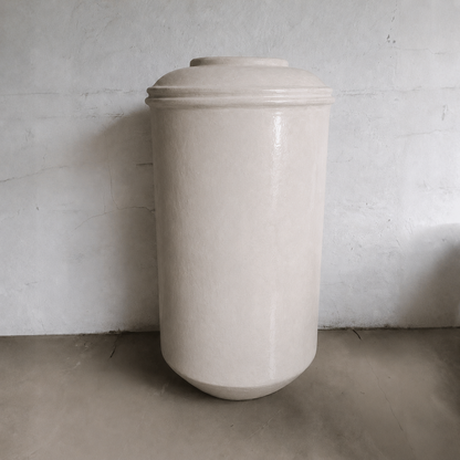 The Damascus Organic Fiberglass Pot - Earth Range by Florastyle By Hingham, a tall cylindrical planter in smooth, off-white finish, stands against a plain wall and concrete floor in a minimalist setting.