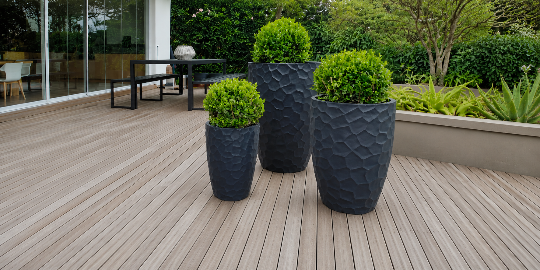 Three textured black planters with green plants on a wooden deck. Japi Prisma Conic Lead set planters.
