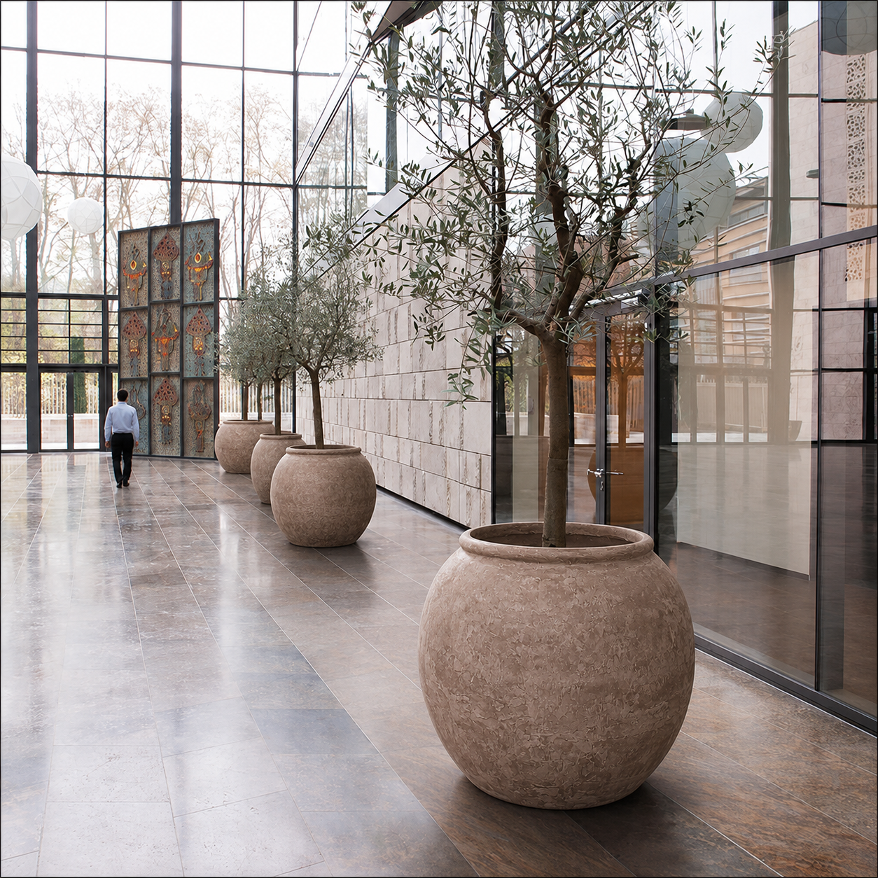A spacious modern hallway with large trees in Florastyle By Hingham’s Delphi Organic Fiberglass Pot - Earth Range, sleek planters, floor-to-ceiling windows, stone tiles, and a single person walking away. Natural light fills the space.