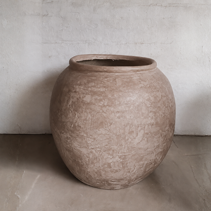 The Florastyle By Hingham Delphi Organic Fiberglass Pot - Earth Range features a large, round, textured unglazed surface in an earthy tone, resting on a smooth floor against a white wall for rustic wabi-sabi appeal.