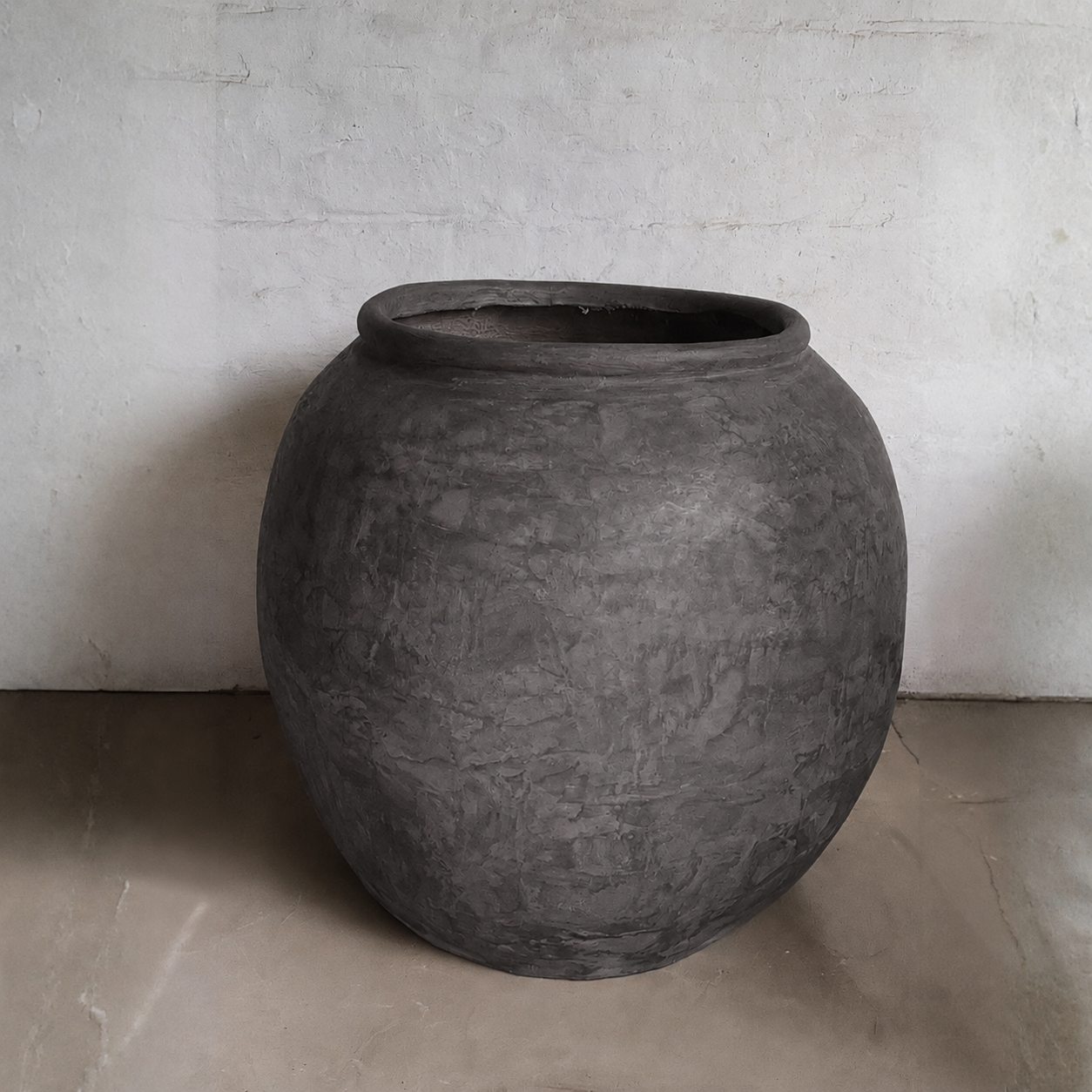 A large, round Delphi Organic Fiberglass Pot - Earth Range by Florastyle By Hingham sits on a light floor against a white textured wall, showcasing wabi-sabi style.