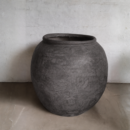 A large, round Delphi Organic Fiberglass Pot - Earth Range by Florastyle By Hingham sits on a light floor against a white textured wall, showcasing wabi-sabi style.