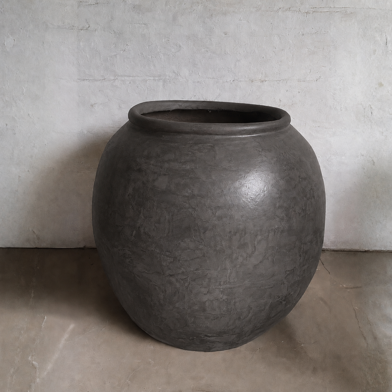 The Delphi Organic Fiberglass Pot - Earth Range by Florastyle By Hingham is a large, round dark gray planter with a wide opening and rustic, slightly uneven surface, shown on a smooth light floor against a textured off-white wall.