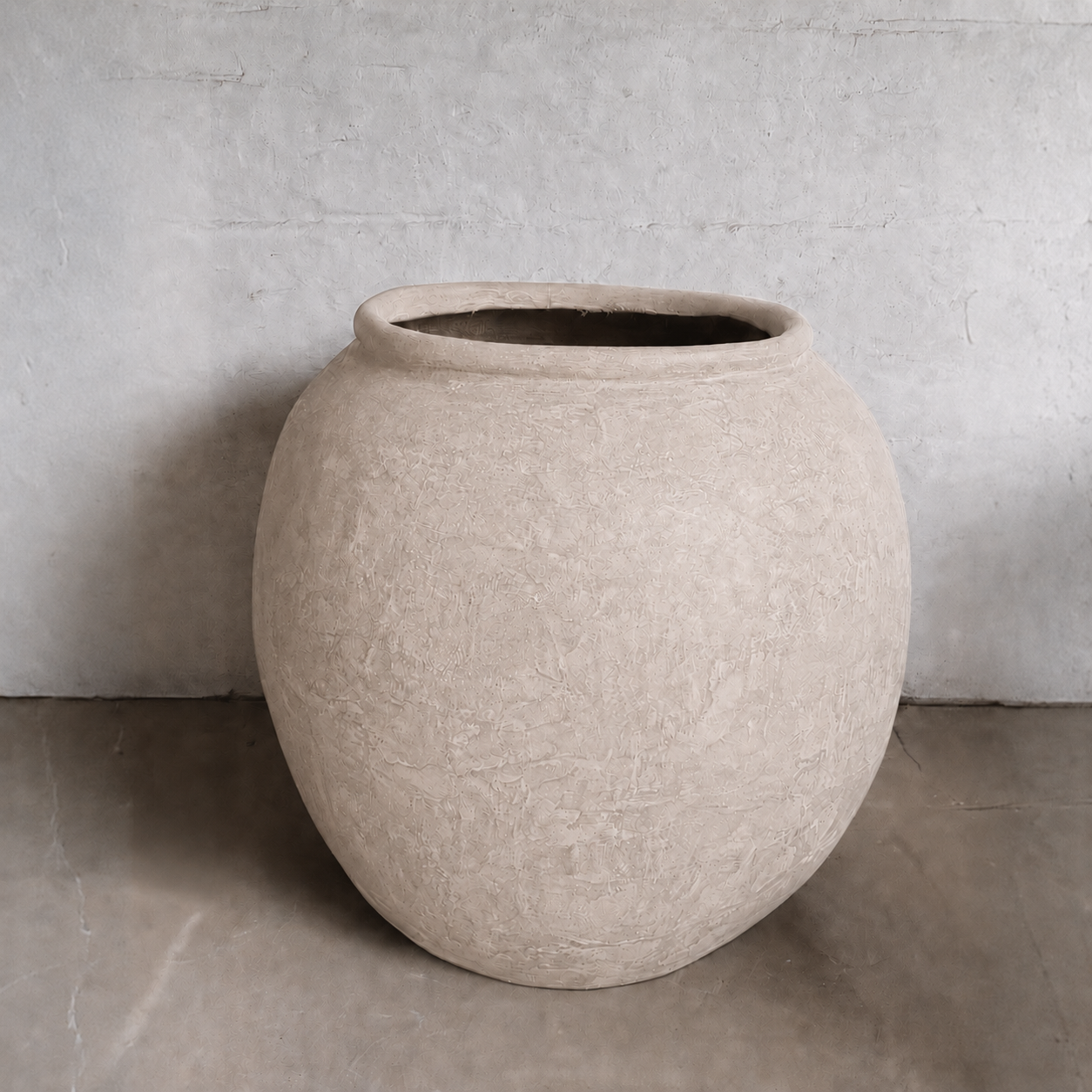 The Delphi Organic Fiberglass Pot - Earth Range by Florastyle By Hingham, with its large round shape and beige finish, sits on a smooth concrete floor against a light gray textured wall, adding subtle elegance to the space.