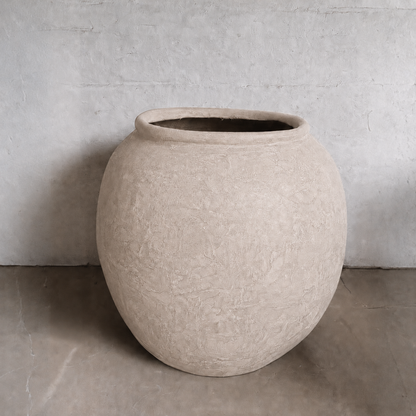 The Delphi Organic Fiberglass Pot - Earth Range by Florastyle By Hingham, with its large round shape and beige finish, sits on a smooth concrete floor against a light gray textured wall, adding subtle elegance to the space.