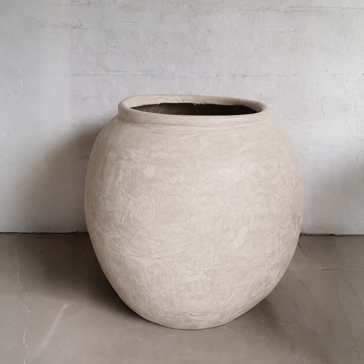The Florastyle By Hingham Delphi Organic Fiberglass Pot - Earth Range is a large, round beige pot with a wide opening and a textured, natural finish. It rests on a smooth floor beside a plain, light wall, featuring no visible decorations.