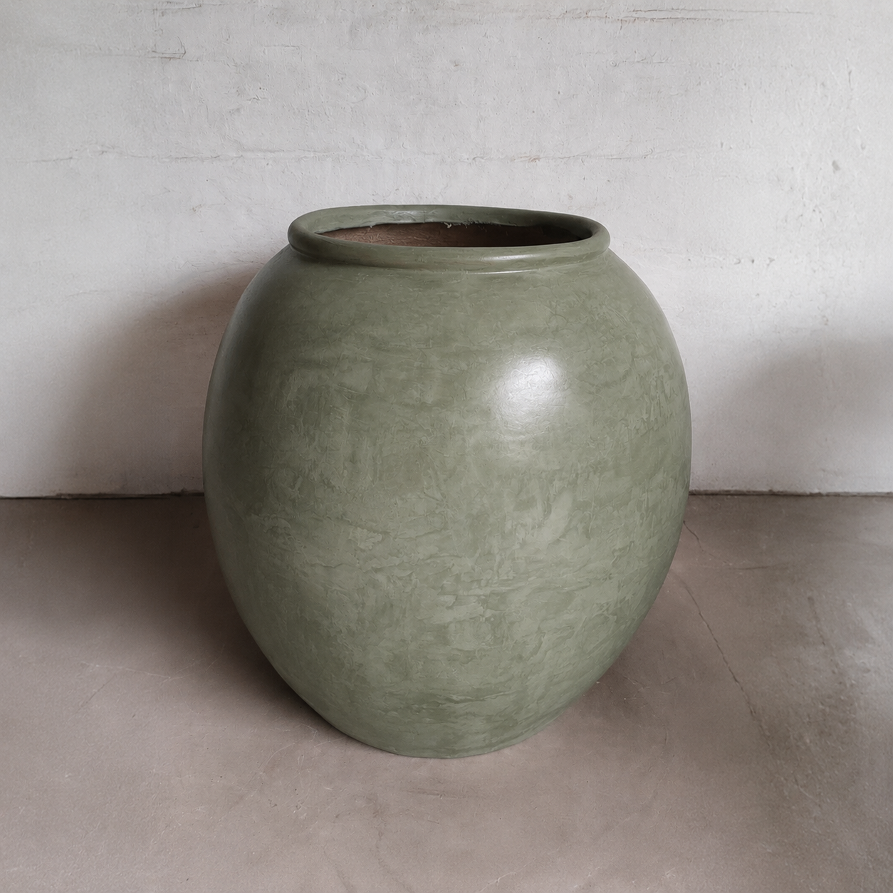 A large, round Florastyle By Hingham Delphi Organic Fiberglass Pot - Earth Range in Vogue Green sits on a smooth, light floor against a plain, white textured wall.