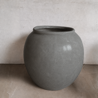 The Delphi Organic Fiberglass Pot - Earth Range by Florastyle By Hingham, featuring a large, round gray design with a wide opening, is placed on a beige floor against a plain light wall.