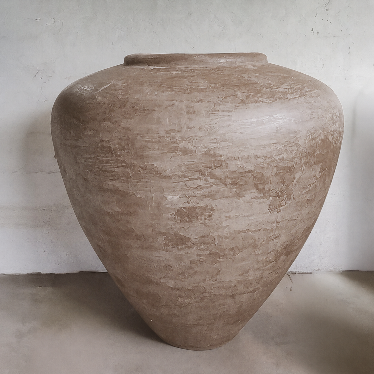 The Mykonos Organic Fiberglass Pot - Earth Range by Florastyle By Hingham, a large, textured brown handcrafted planter with a wide body and narrow base, adds sculptural décor against a light wall on a smooth floor.