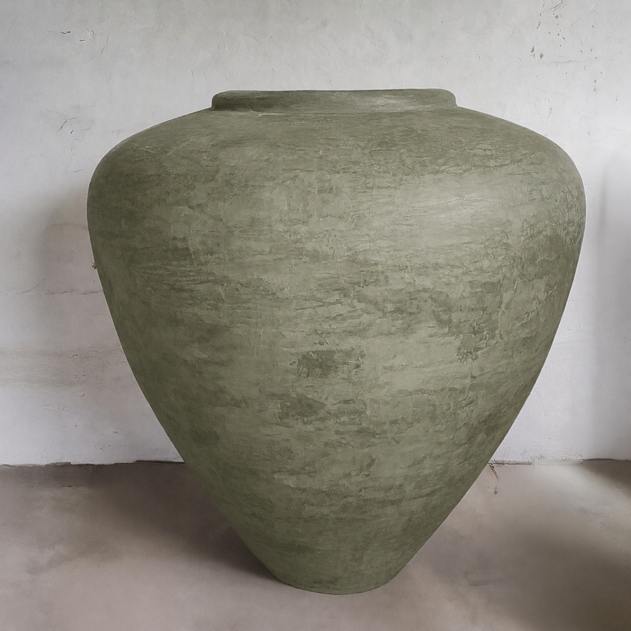 The Mykonos Organic Fiberglass Pot from Florastyle By Hingham's Earth Range features a large, textured, olive-green design with a rounded body and narrow opening, set against a plain light wall on a smooth floor.