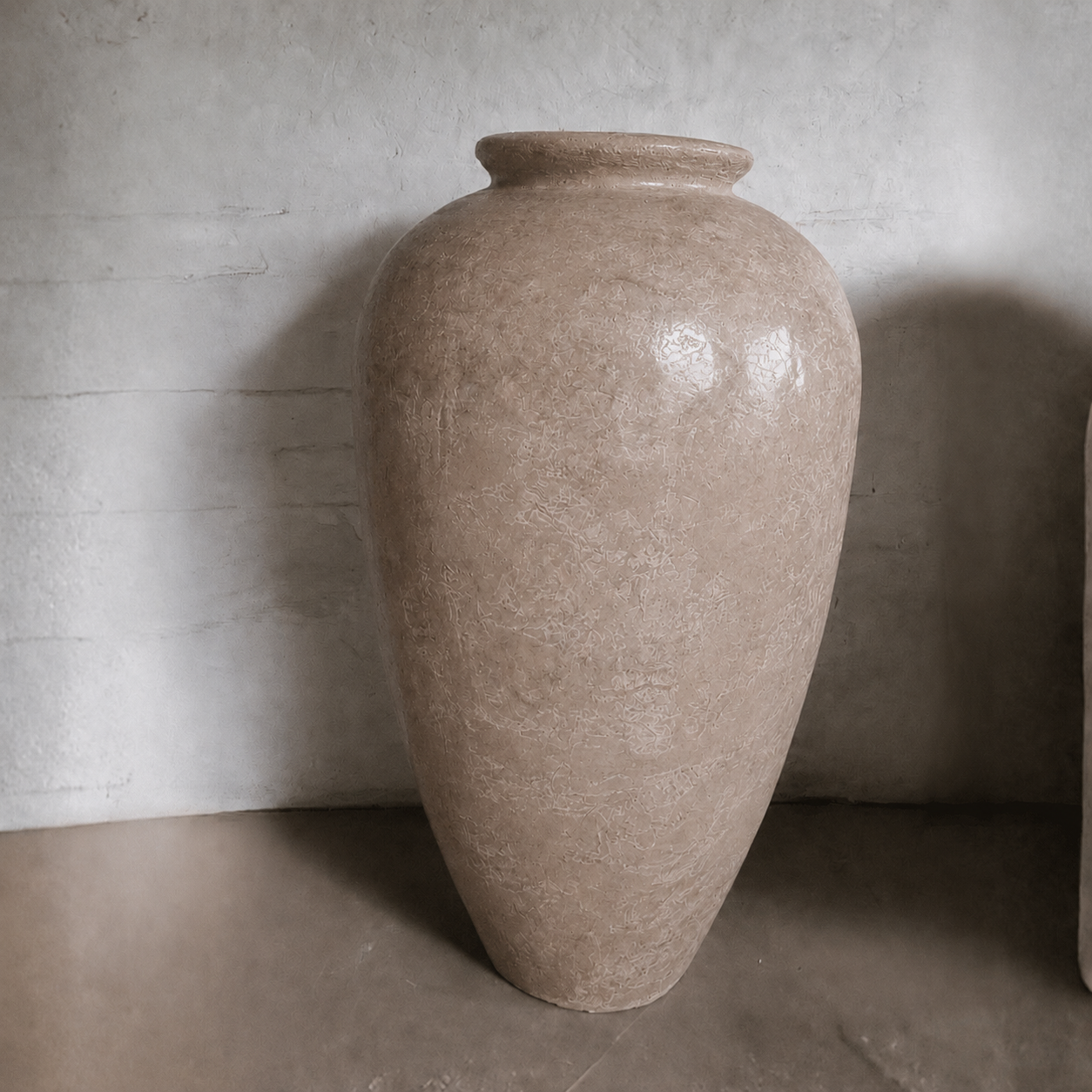 The Rhodes Organic Fiberglass Pot from Florastyle By Hingham’s Earth Range, featuring a wide body and narrow, flared rim in beige, stands on a smooth floor against a textured, light-colored wall.