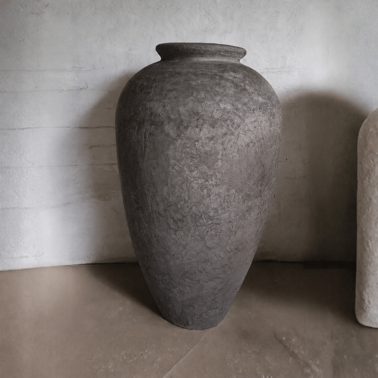 The large, textured Rhodes Organic Fiberglass Pot—Earth Range by Florastyle By Hingham features a wide opening and simple, rounded design. It stands on a smooth floor against a light, slightly textured wall and appears handmade.