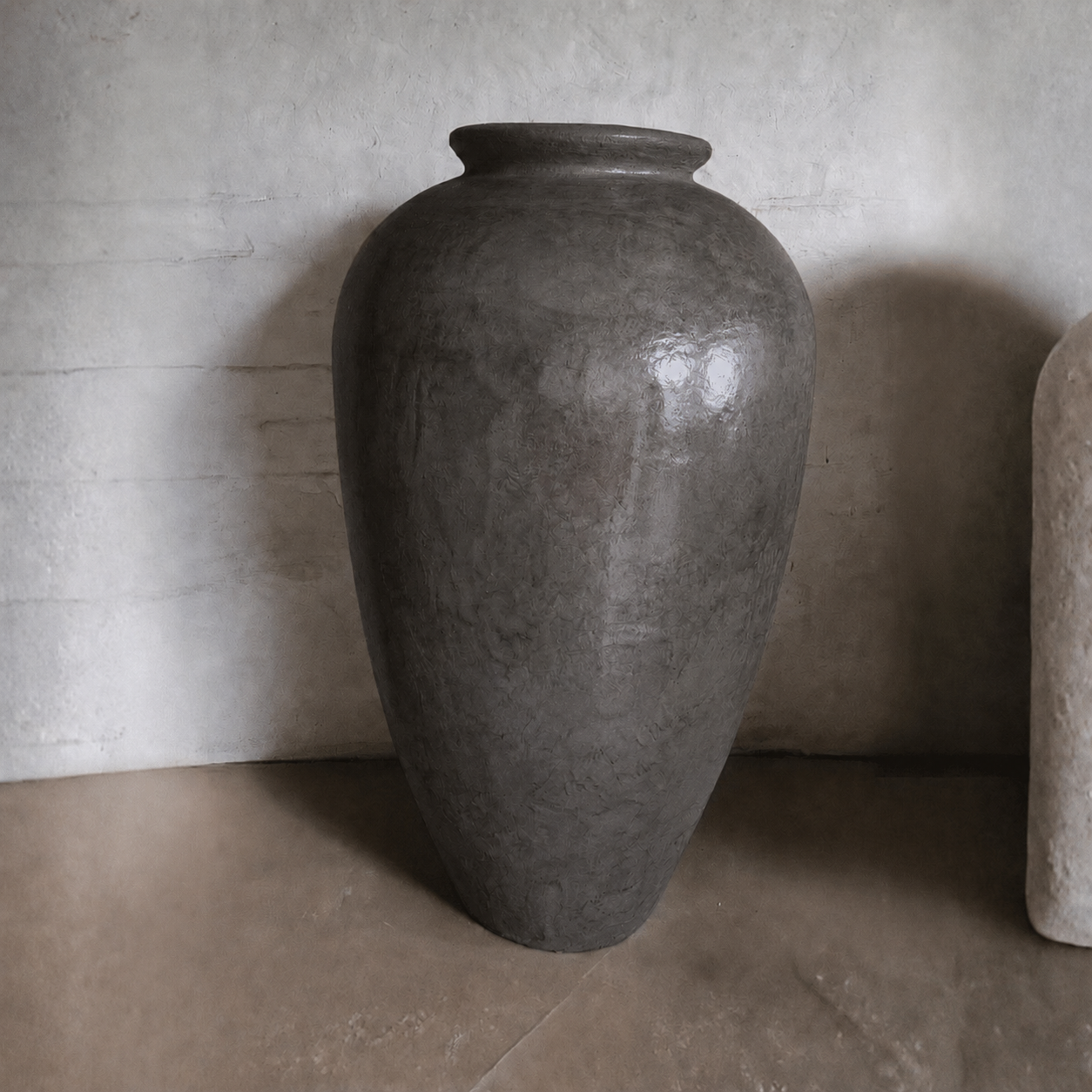 A large, dark gray Rhodes Organic Fiberglass Pot - Earth Range by Florastyle By Hingham, featuring a rounded body and narrow neck with a smooth, slightly reflective finish, sits on a brown surface against a textured light wall.