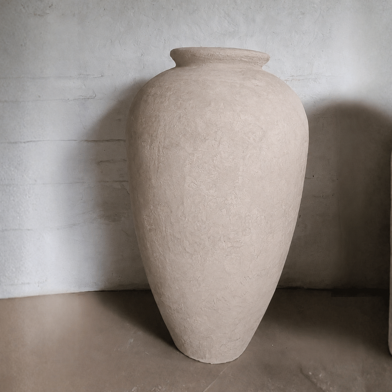 The Rhodes Organic Fiberglass Pot - Earth Range by Florastyle By Hingham, featuring a large beige textured finish with a wide body and narrow opening, stands on a smooth floor against a light-colored textured wall.