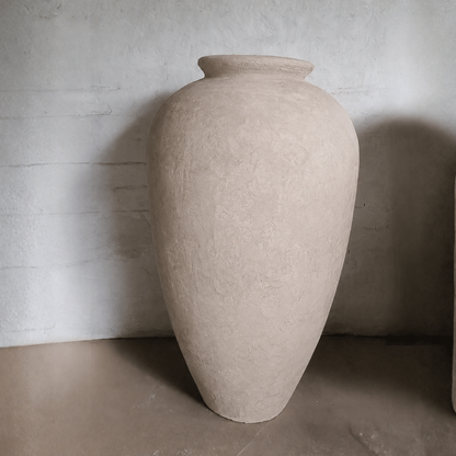 The Rhodes Organic Fiberglass Pot - Earth Range by Florastyle By Hingham, featuring a large beige textured finish with a wide body and narrow opening, stands on a smooth floor against a light-colored textured wall.