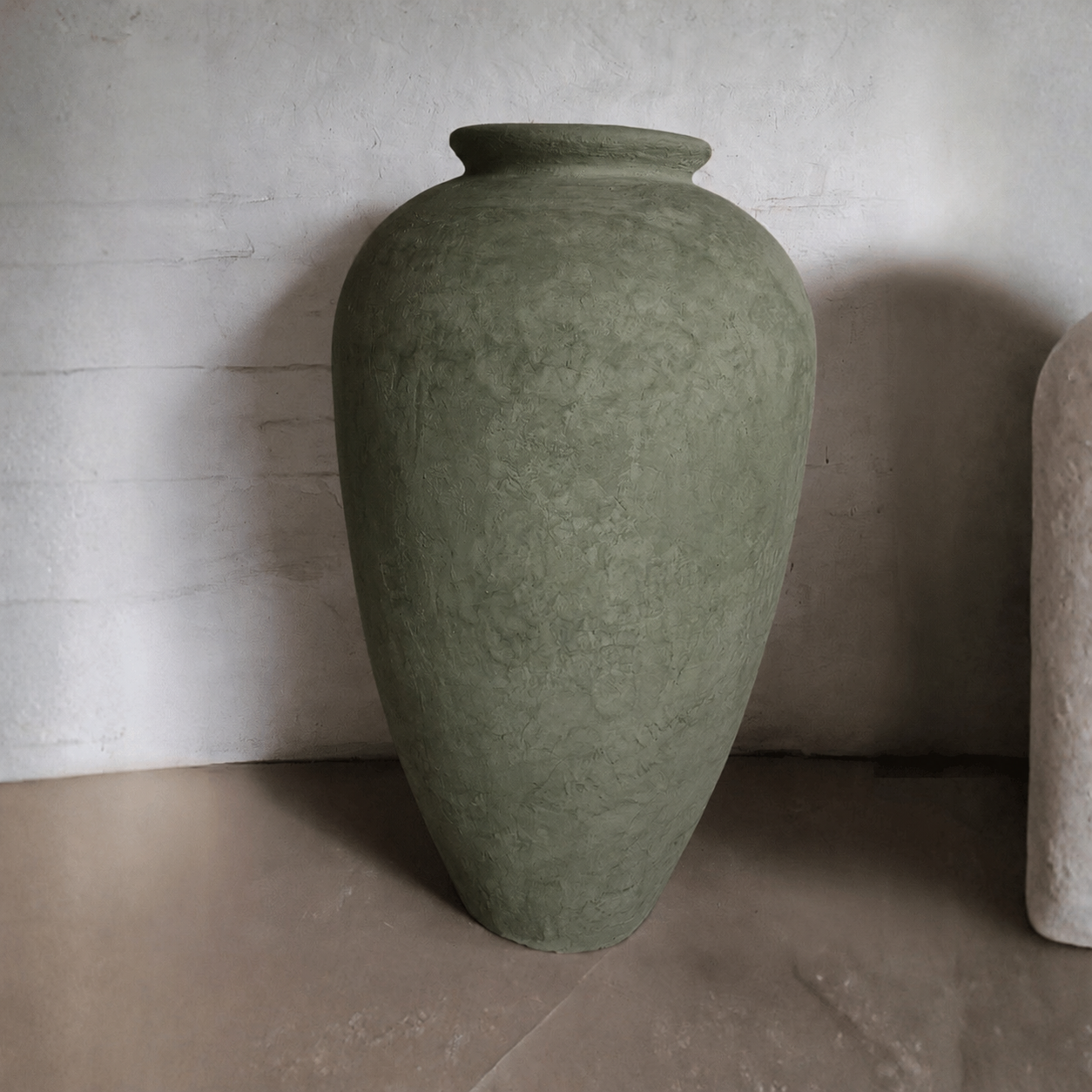A Rhodes Organic Fiberglass Pot - Earth Range by Florastyle By Hingham, large and textured in olive green, with a wide body and narrow opening, sits on a neutral floor against a light, subtly textured wall.