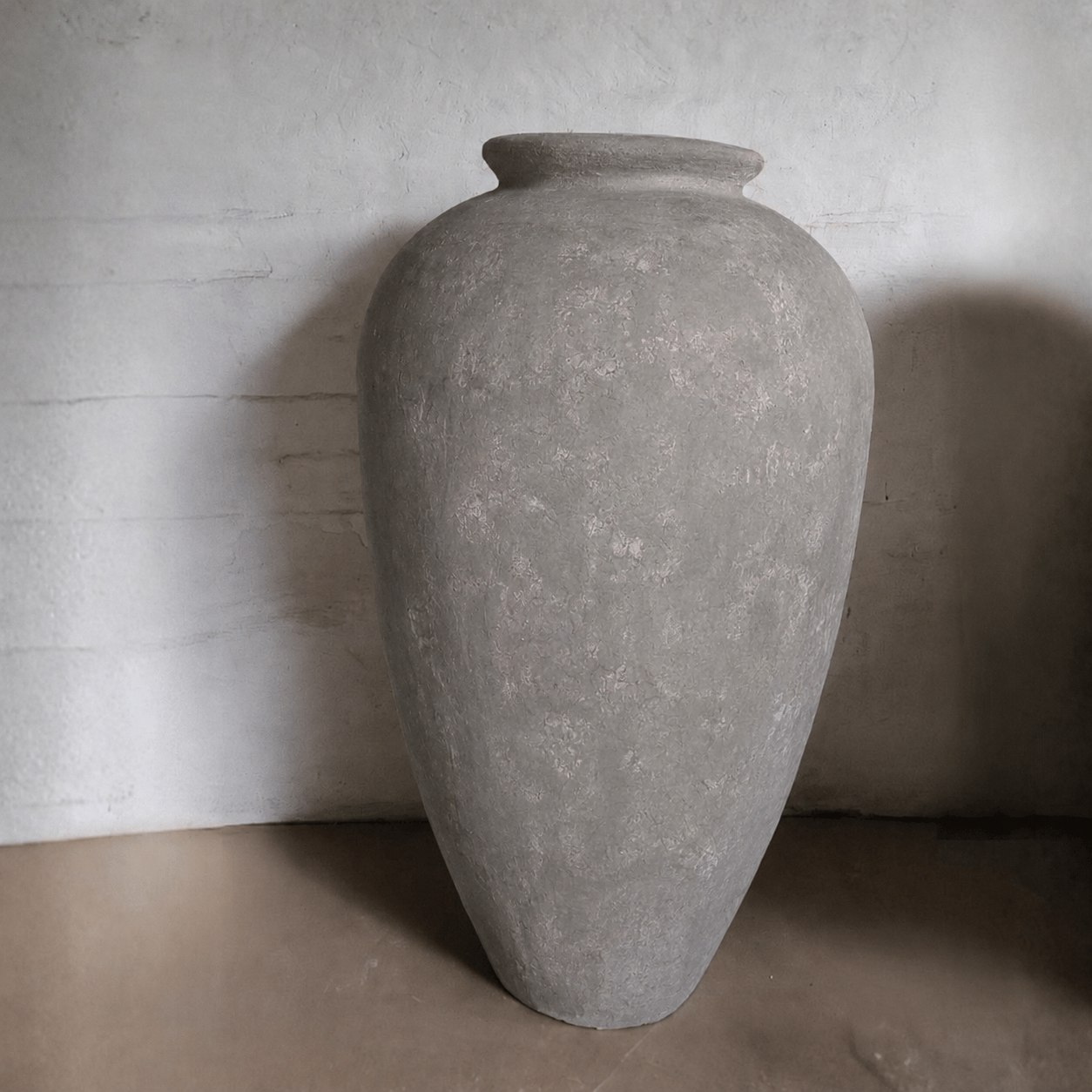 A large Rhodes Organic Fiberglass Pot - Earth Range by Florastyle By Hingham sits on a brown floor against a textured white wall. Its wide rim and weathered, gray finish are highlighted by shadows on the left side, emphasizing its rustic look.