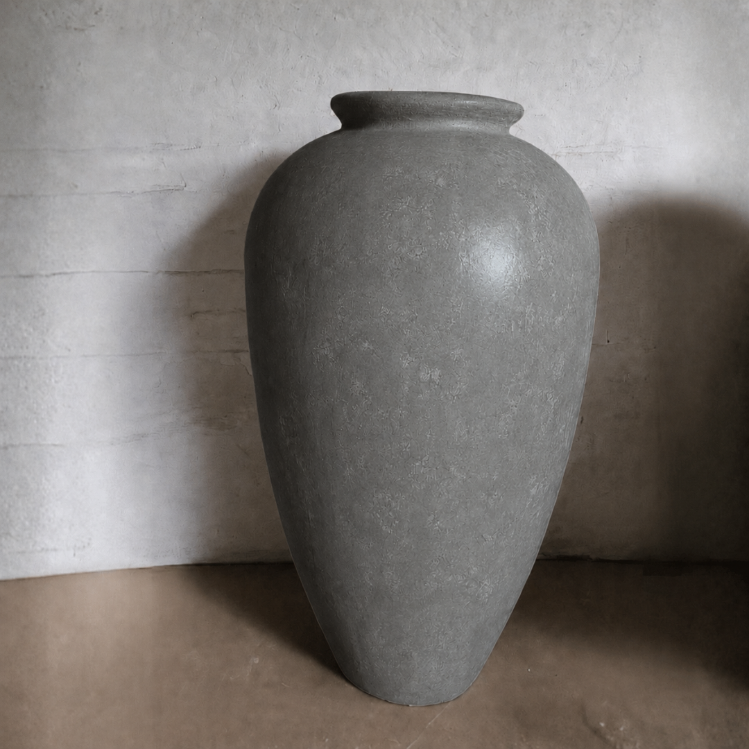 The Rhodes Organic Fiberglass Pot - Earth Range by Florastyle By Hingham, a large gray matte planter with a narrow opening, sits on a brown surface against a textured light-colored wall.