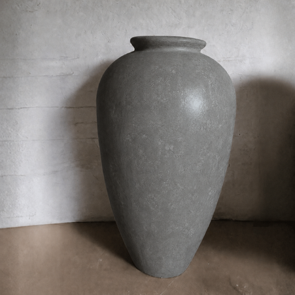 The Rhodes Organic Fiberglass Pot - Earth Range by Florastyle By Hingham, a large gray matte planter with a narrow opening, sits on a brown surface against a textured light-colored wall.