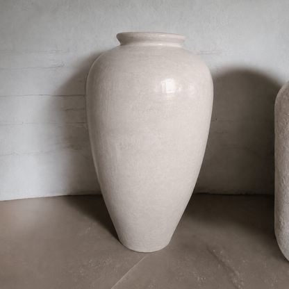 The Rhodes Organic Fiberglass Pot - Earth Range by Florastyle By Hingham, featuring a large off-white rounded body and narrow neck, stands on a smooth floor against a textured light-gray wall.
