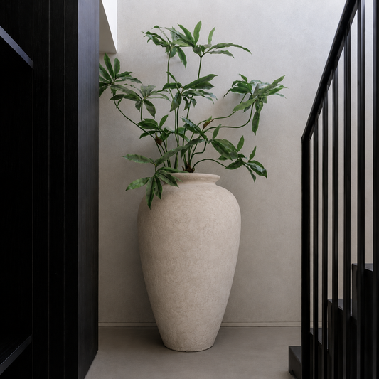 A large beige Rhodes Organic Fiberglass Pot - Earth Range by Florastyle By Hingham, filled with green leafy branches, stands in a minimalist corner beside a black staircase and a smooth neutral wall.