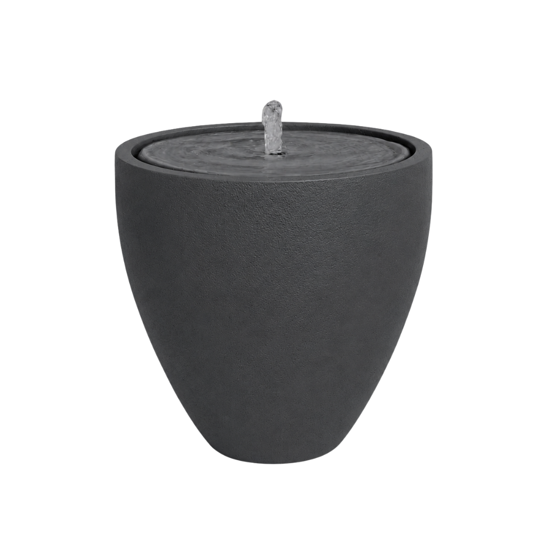 The Florastyle By Hingham Water Feature - Rustic Ellipse Small is a modern, dark gray, round tabletop fountain with a smooth surface. Water gently bubbles from the central spout and flows over the flat top. Background is plain white.