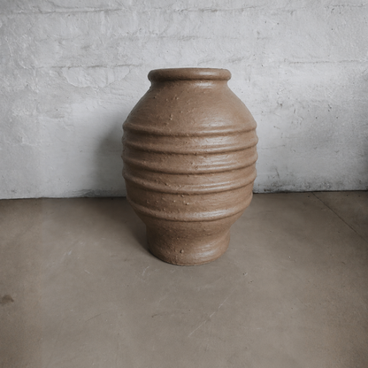 A large Sparta Organic Fiberglass Pot from Florastyle By Hingham, in an earth-tone finish with horizontal ridges, sits on a gray concrete floor against a white, rough brick wall.