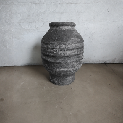 The Sparta Organic Fiberglass Pot - Earth Range by Florastyle By Hingham, in gray with horizontal ridges, stands on a bare concrete floor against a white textured wall—ideal for organic modern decor.