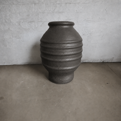 A large, dark Sparta Organic Fiberglass Pot from Florastyle By Hingham's Earth Range, featuring a wide body and narrow neck, stands on a concrete floor against a plain, textured white wall.