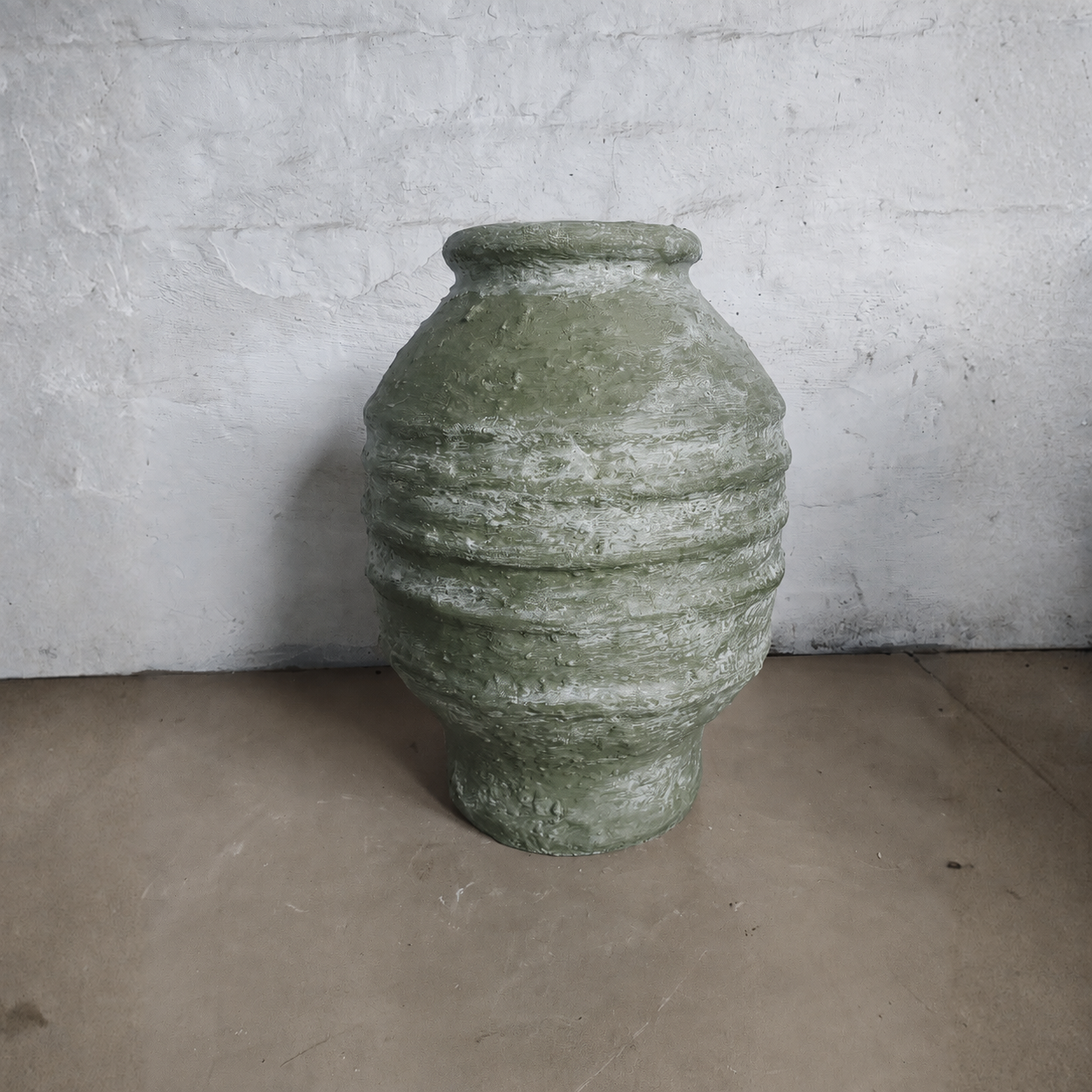The Sparta Organic Fiberglass Pot - Earth Range by Florastyle By Hingham, featuring a large textured green design with horizontal ridges, sits on a concrete floor against a white wall—ideal for organic modern decor.