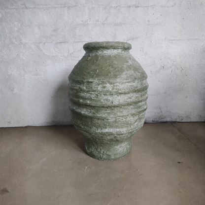 The Sparta Organic Fiberglass Pot - Earth Range by Florastyle By Hingham, featuring a large textured green design with horizontal ridges, sits on a concrete floor against a white wall—ideal for organic modern decor.