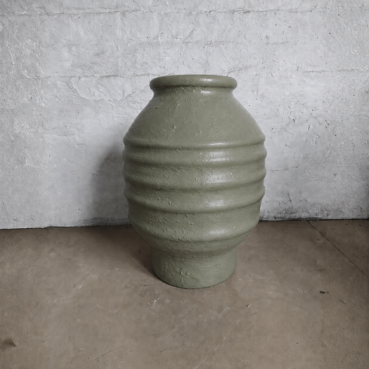 A large, textured olive-green Sparta Organic Fiberglass Pot from Florastyle By Hingham's Earth Range, featuring horizontal ridges, is placed against a rough white wall on a plain light brown floor.