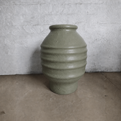 A large, textured olive-green Sparta Organic Fiberglass Pot from Florastyle By Hingham's Earth Range, featuring horizontal ridges, is placed against a rough white wall on a plain light brown floor.
