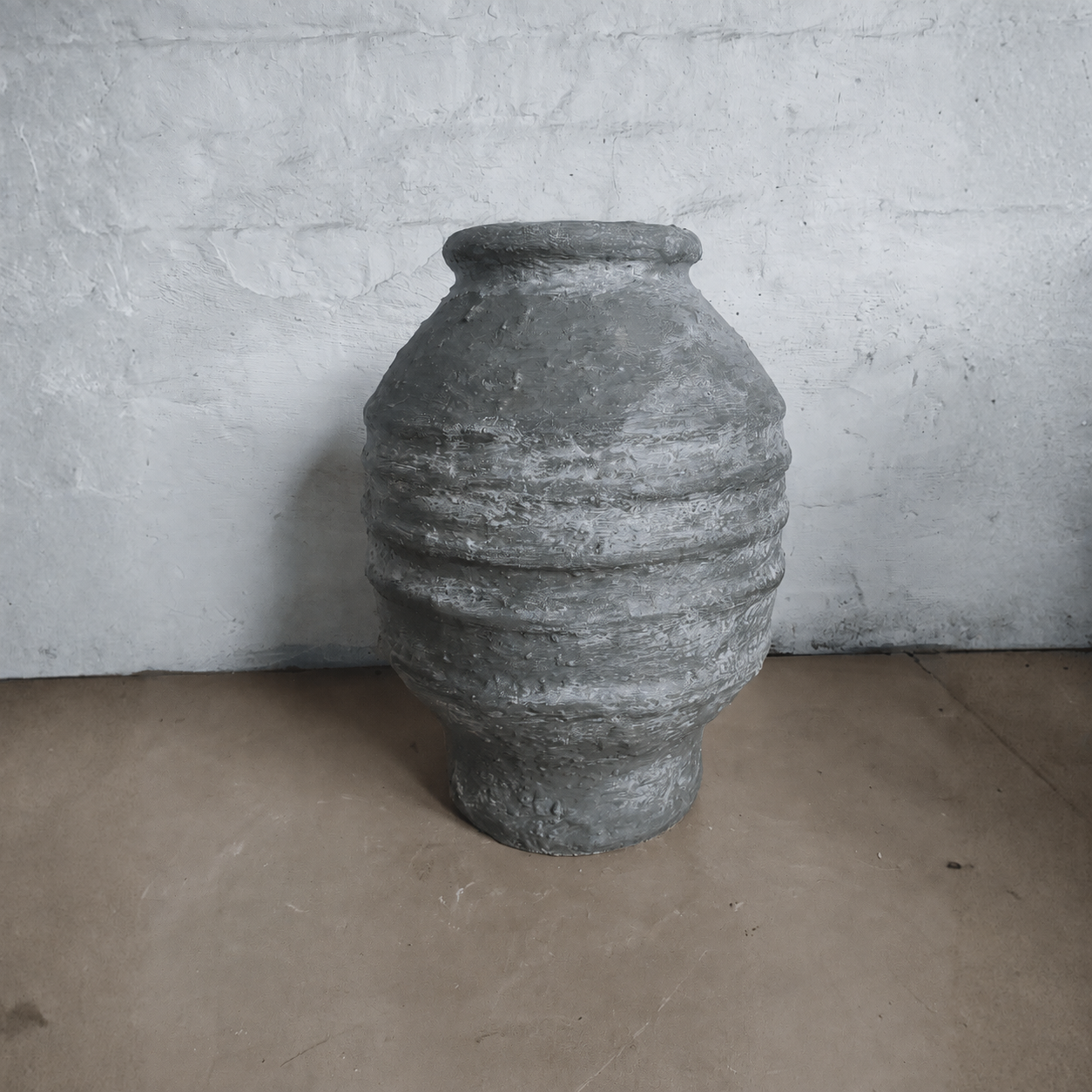 A large Sparta Organic Fiberglass Pot - Earth Range by Florastyle By Hingham, featuring textured horizontal ridges in gray, sits on a brown floor against a white stone wall, adding an organic modern touch.