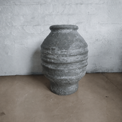 A large Sparta Organic Fiberglass Pot - Earth Range by Florastyle By Hingham, featuring textured horizontal ridges in gray, sits on a brown floor against a white stone wall, adding an organic modern touch.