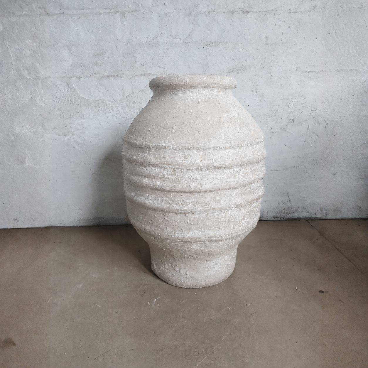 The Sparta Organic Fiberglass Pot - Earth Range by Florastyle By Hingham is a large, textured beige planter with horizontal ridges—an ideal lightweight accent for organic modern decor on a bare concrete floor.