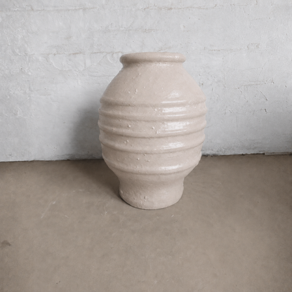A large, rounded Sparta Organic Fiberglass Pot - Earth Range by Florastyle By Hingham, featuring horizontal ridges and a cream finish, is placed on a beige floor against a textured white wall.
