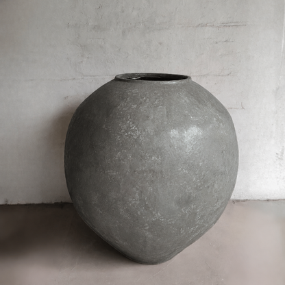 The Syracuse Organic Fiberglass Pot – Earth Range by Florastyle By Hingham is a large, round gray planter with a weathered texture, wide body, and narrow opening. It sits against a plain, light wall on a smooth floor.