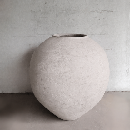 The Syracuse Organic Fiberglass Pot from the Earth Range by Florastyle By Hingham, featuring a large, rounded design with a narrow opening, rests on a smooth concrete floor against a plain light gray wall.