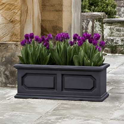 The Classic Versailles Japi Window Box by Florastyle By Hingham, in black, showcases blooming purple tulips and sits on a stone patio beside textured stone walls, providing a stylish indoor or outdoor planter option.