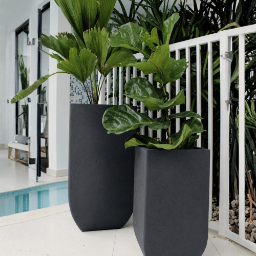 Two Florastyle By Hingham Verticale Square Japi Planters with lush green indoor plants sit by a white railing next to a pool in a modern, bright room with glass doors and tropical decor.